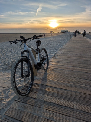 Ein Fahrrad steht auf einem hölzernen Steg am Strand bei Sonnenuntergang; im Hintergrund laufen ein paar Menschen am Meer entlang.