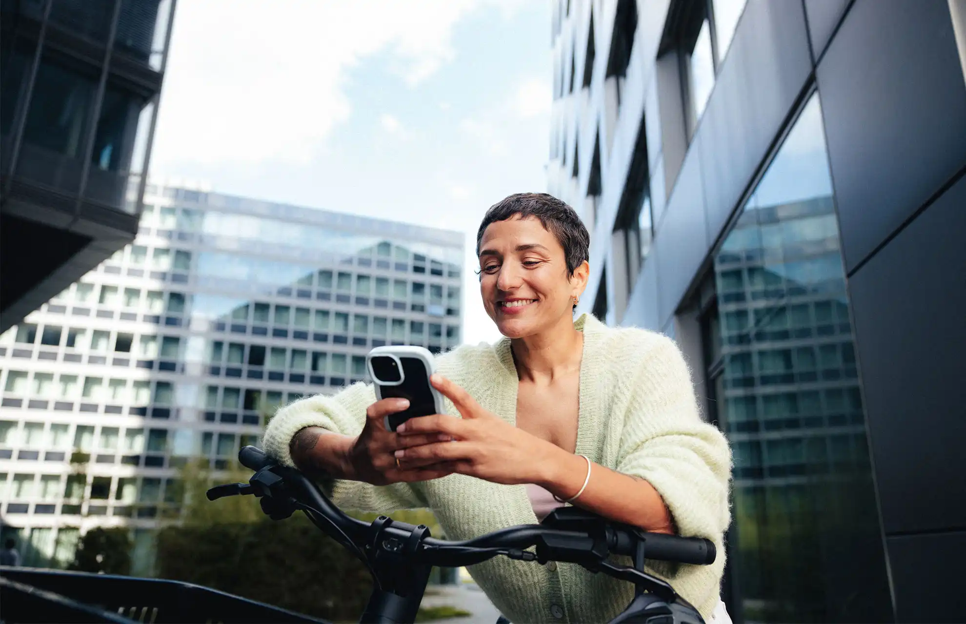 Chefin lehnt vor einem Bürogebäude auf ihrem Dienstrad und schaut lächelnd auf ihr Handy