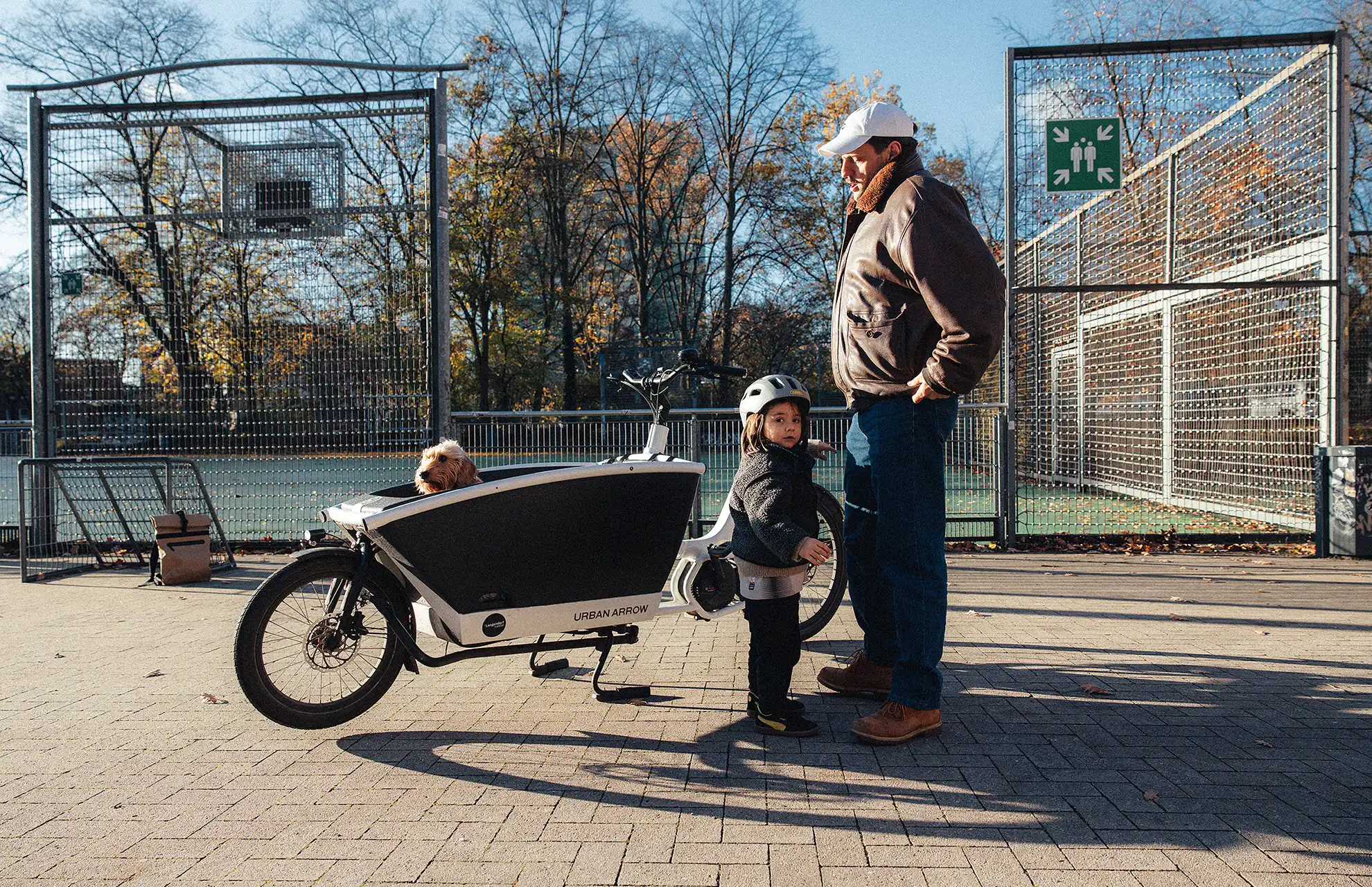 Vater und Tochter stehen neben dem Lastenrad. Tochter mit Helm schaut in die Kamera, Vater schaut aufs Kind. Vorne im Lastenrad schaut ein kleiner Hund raus und blickt unschlüssig.