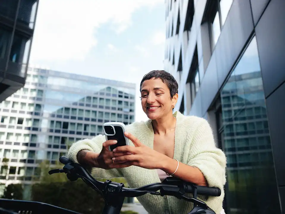 Chefin lehnt vor einem Bürogebäude auf ihrem Dienstrad und schaut lächelnd auf ihr Handy